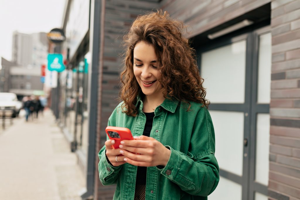 outdoor-portrait-lovely-stylish-girl-with-curls-wearing-green-shirt-using-smartphone-with-smile-carefree-young-caucasian-girl-is-using-modern-smartphone-standing-outdoors